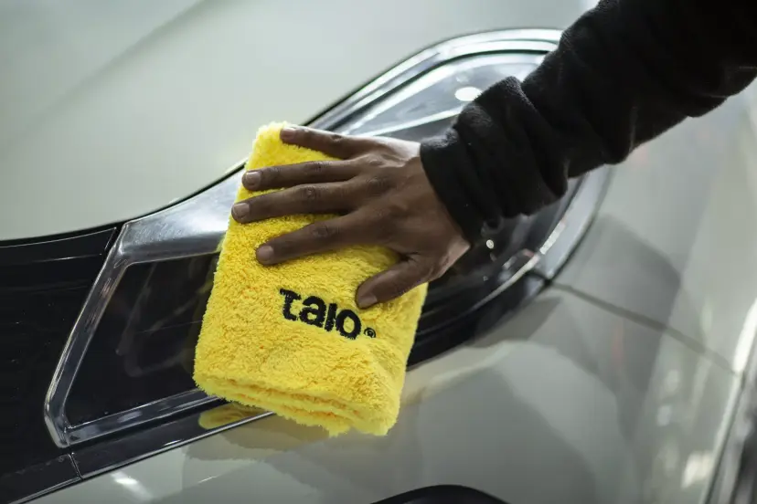 Hand holding a yellow Talo towel against the headlight of a white car